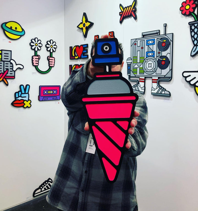 photograph of a man holding an artwork by nick chaffe titled spray me if you can. the artwork resembles an ice cream cone in colours pink and grey, with a spray button atop in blue