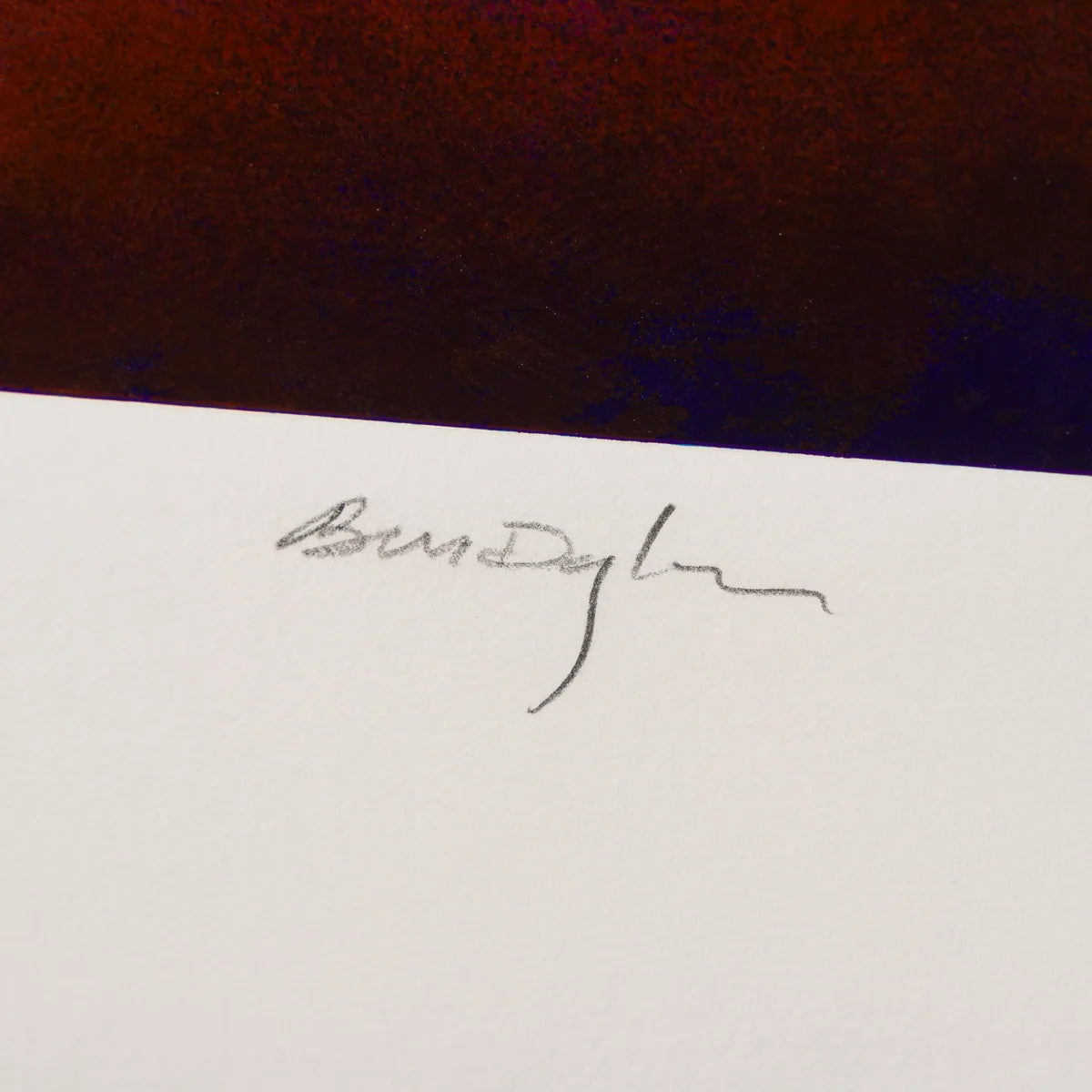 A close-up of a white card or paper with a small black handwritten signature, set against a dark reddish-brown background which is Bob Dylan's signature on one of his editioned prints