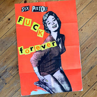 An image of the Jamie Reid artwork 'Fuck Forever', laid on a wooden floor. The artwork version has a red background, and lettering stating Sex Pistols Fuck Forever. 