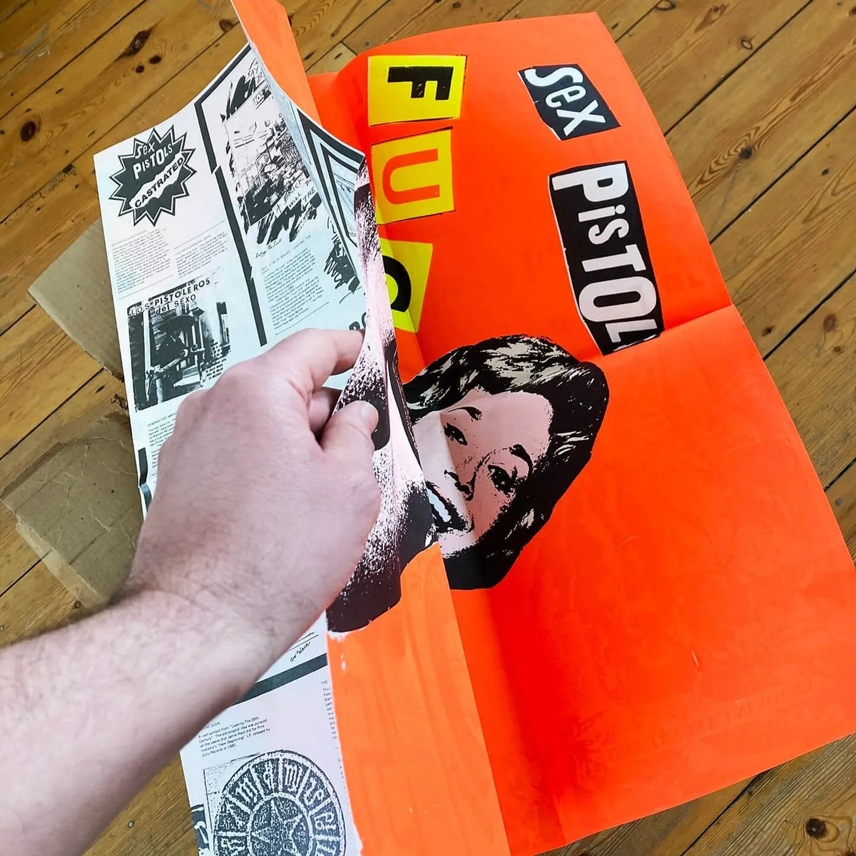 An image of the Jamie Reid artwork 'Fuck Forever', being folded by a hand to show a printed back cover of the artwork, against a wooden floor. The artwork version has a red background, and lettering stating Sex Pistols Fuck Forever. 