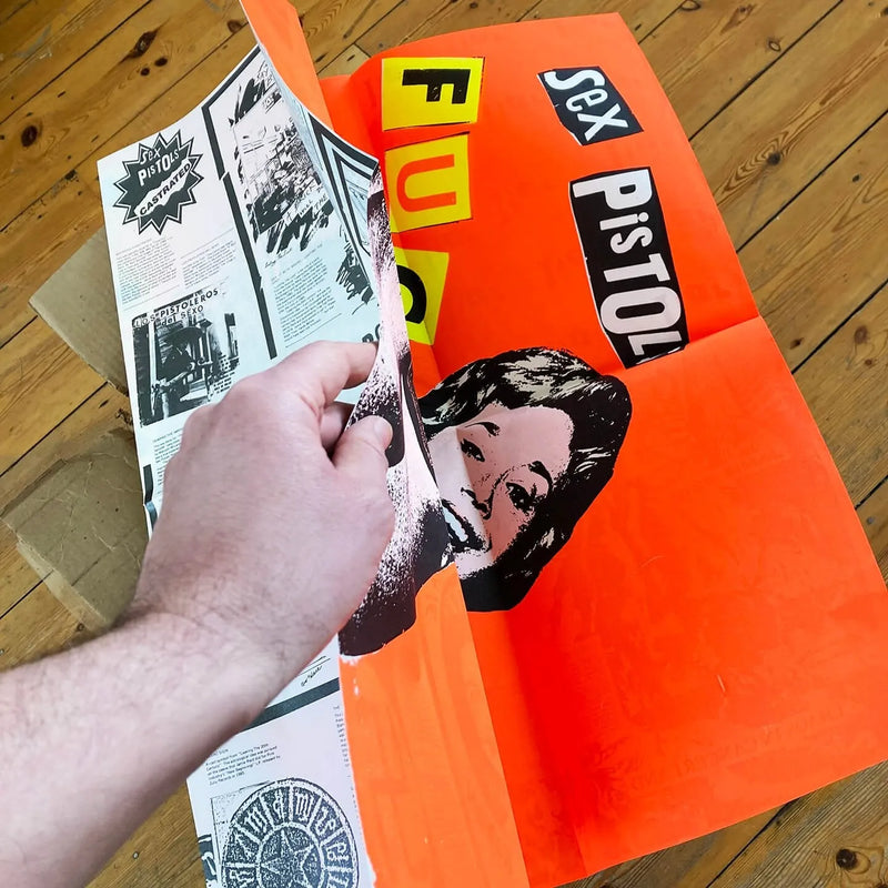 An image of the Jamie Reid artwork 'Fuck Forever', being folded by a hand to show a printed back cover of the artwork, against a wooden floor. The artwork version has a red background, and lettering stating Sex Pistols Fuck Forever. 