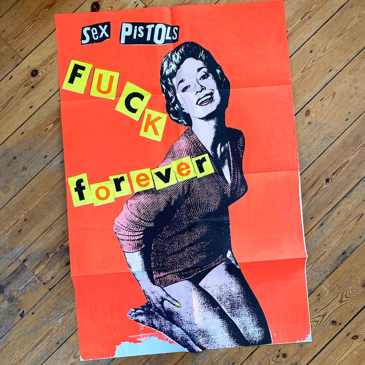 An image of the Jamie Reid artwork 'Fuck Forever', laid on a wooden floor. The artwork version has a red background, and lettering stating Sex Pistols Fuck Forever. 