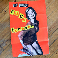 An image of the Jamie Reid artwork 'Fuck Forever', laid on a wooden floor. The artwork version has a red background, and lettering stating Sex Pistols Fuck Forever. 