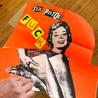 A zoomed in image of the Jamie Reid artwork 'Fuck Forever', being held by a hand, with the upper part of the poster against a wooden floor. The artwork version has a red background, and lettering stating Sex Pistols Fuck Forever. 