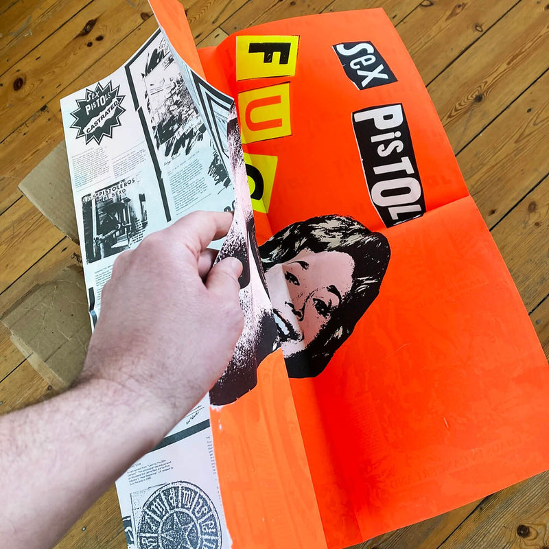An image of the Jamie Reid artwork 'Fuck Forever', being folded by a hand to show a printed back cover of the artwork, against a wooden floor. The artwork version has a red background, and lettering stating Sex Pistols Fuck Forever. 