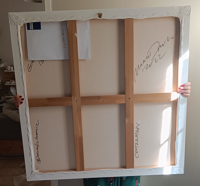 The back side of a framed canvas print, showing the wooden stretcher bars, white hanging surface, and several handwritten signatures, held up by a person in an indoor setting. The artwork is by Mark Drew