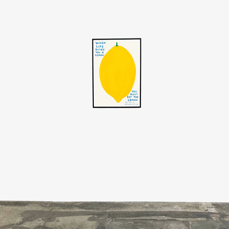 An image of an artwork by David Shrigley titled 'When Life Gives You A Lemon' in a black frame hung on a white wall in a room with a grey floor. The piece features a large, cartoon-like depiction of a lemon, with text at the top right stating 'WHEN LIFE GIVES YOU A LEMON', and in the bottom right continuing 'YOU MUST EAT THE LEMON', 'ALL OF IT', 'INCLUDING THE SKIN'. This is a David Shrigley Poster or Print